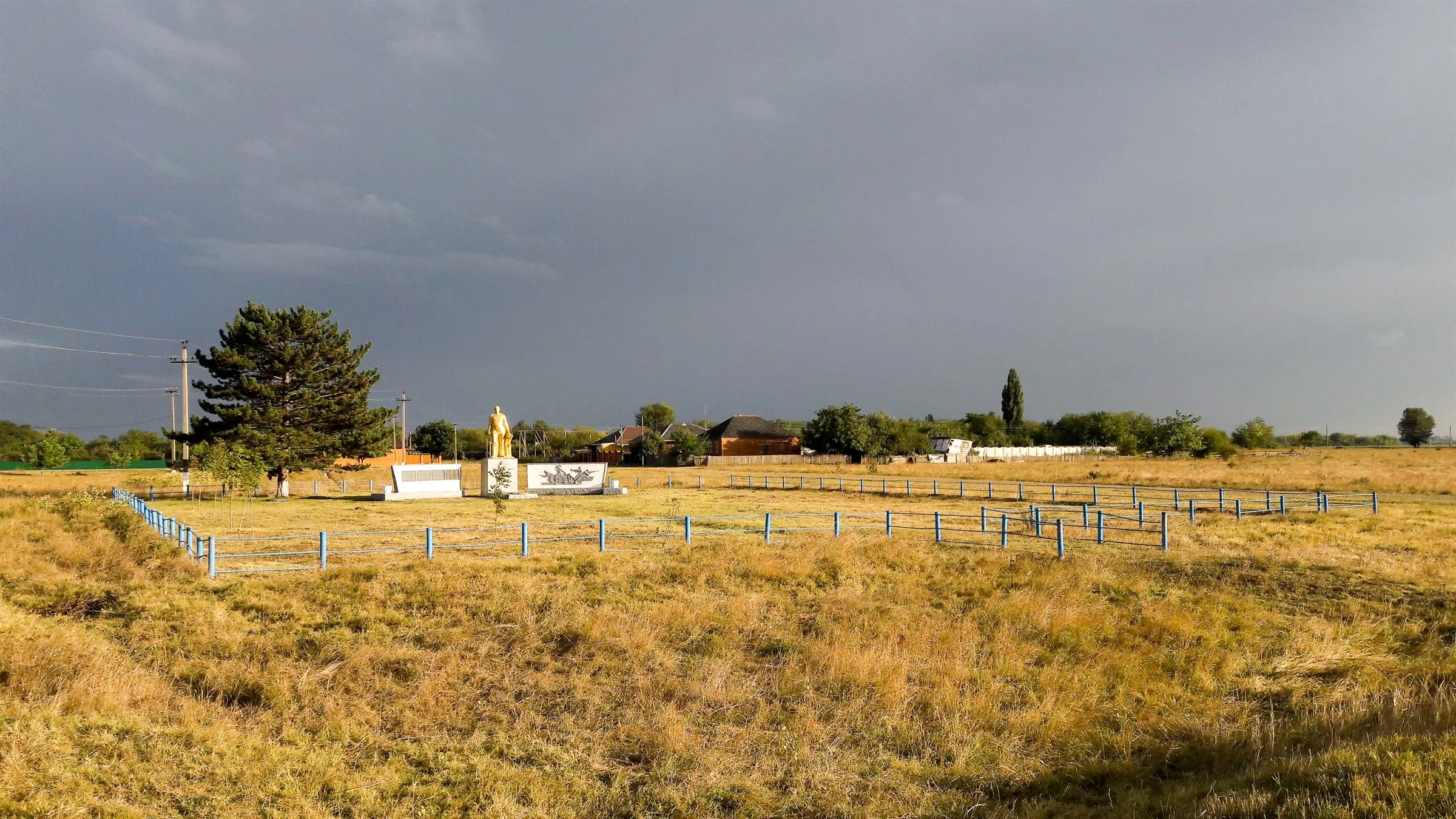 Weites Grasland am Rand eines Dorfes unter dunklem Himmel, die Abendsonne beleuchtet ein kleines Denkmal hinter einem Zaun.
