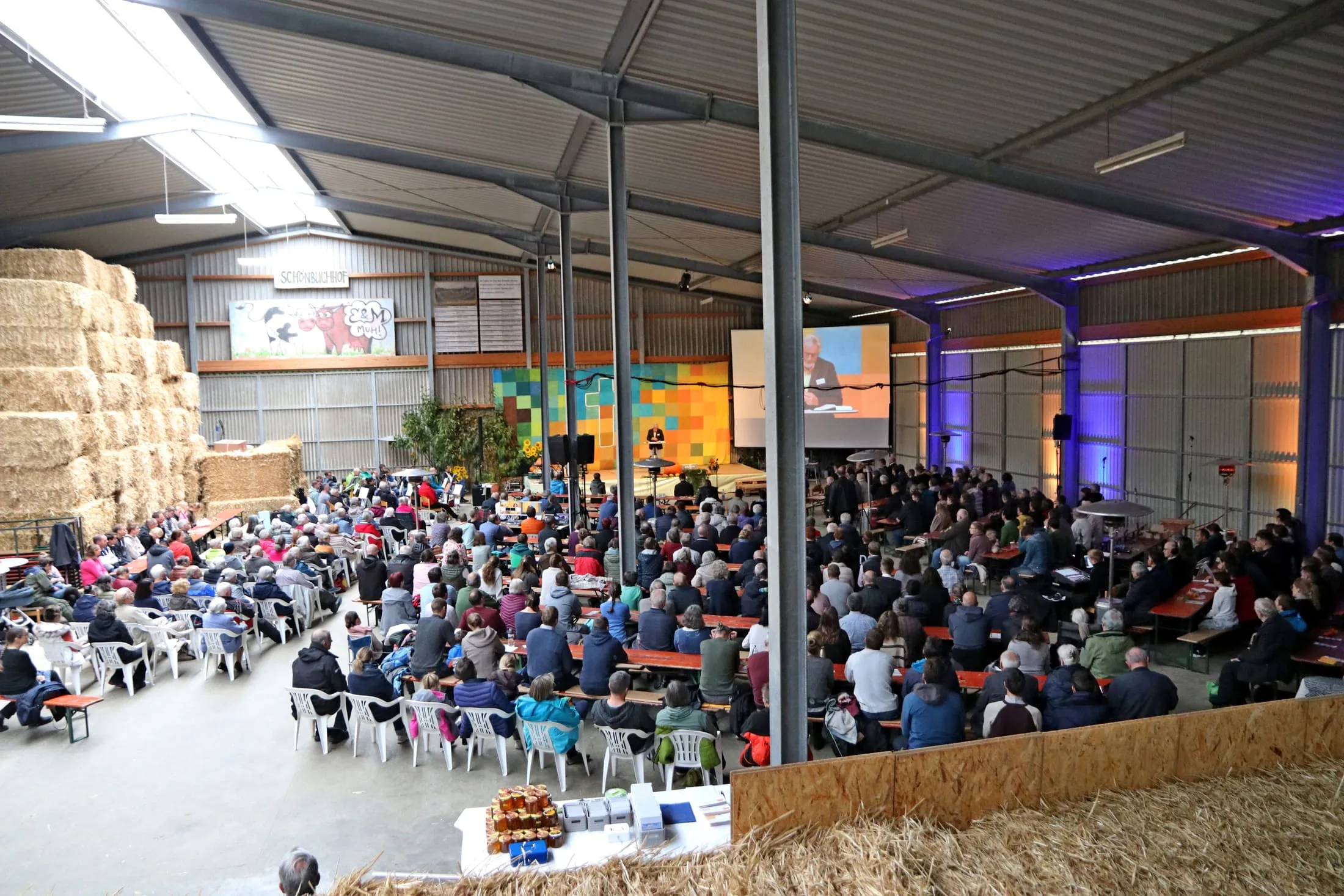 Der volle Saal beim Missionstag: Besucher sitzen an langen Tischen, vorn läuft ein Bericht auf der Leinwand.