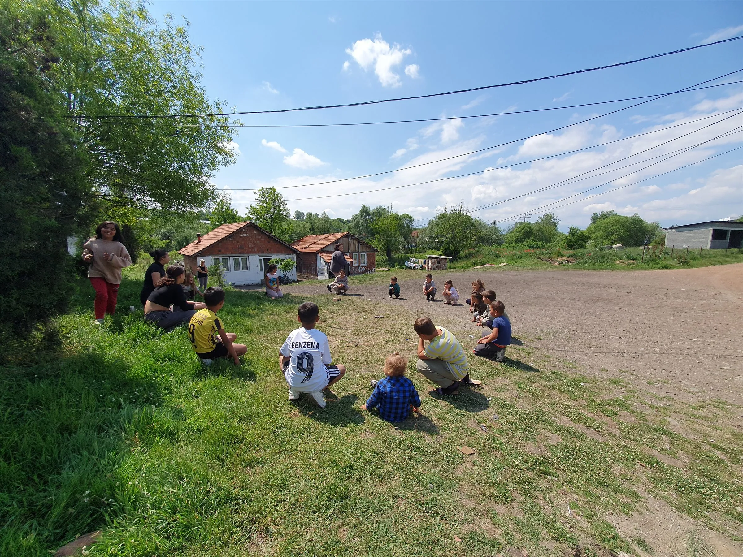 Kinderstunde im Freien im Kosovo: Kinder sitzen im Kreis auf einem Dorfplatz, am Rand sitzen Mitarbeiterinnen im Gras.