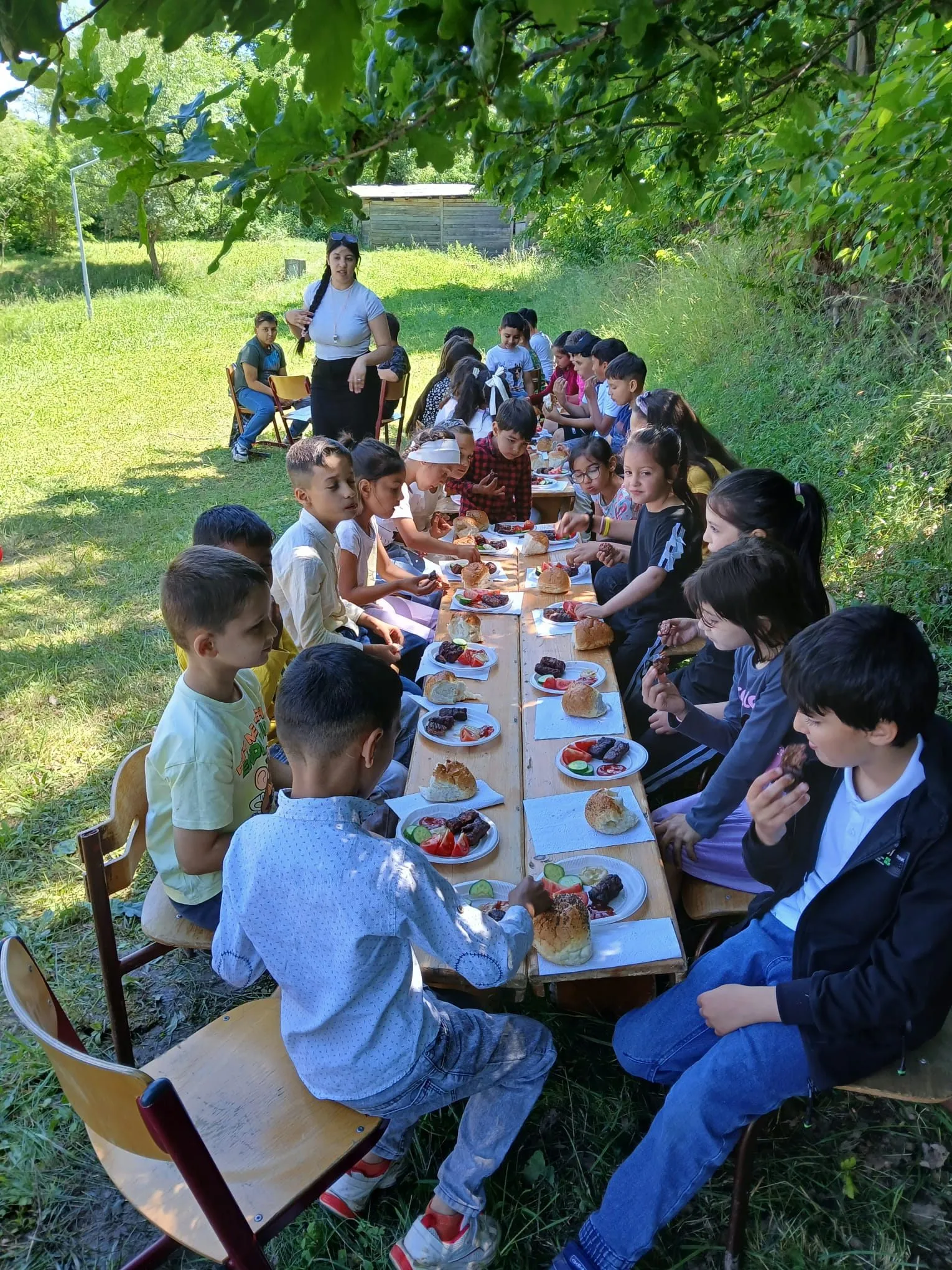 Kinder essen an einer langen Tafel im Freien unter Bäumen.