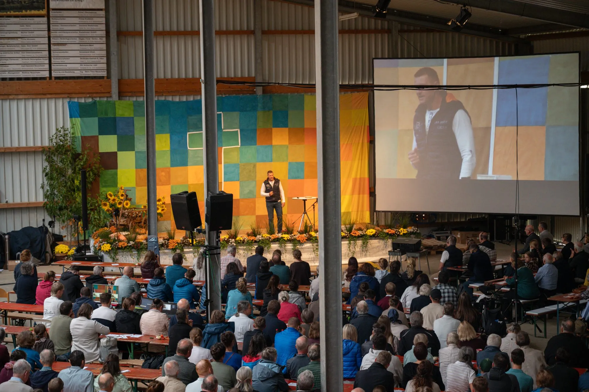 Missionstag 2026: Blick von oben in die volle Halle, vorne spricht ein Redner vor einer bunten Wand, rechts zeigt ihn eine Leinwand.