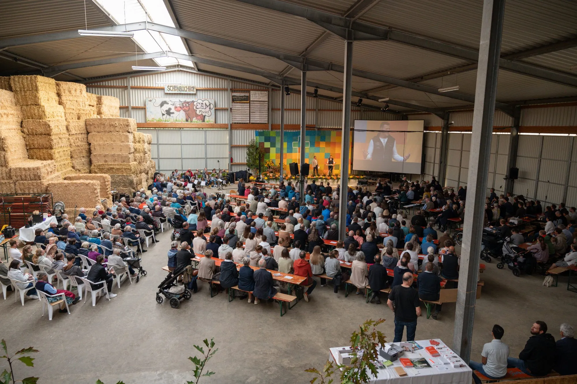 Der Missionstag 2026 von oben: die Halle des Schönbuchhofs ist voll besetzt, vorne die Bühne mit Leinwand, links die gestapelten Strohballen.