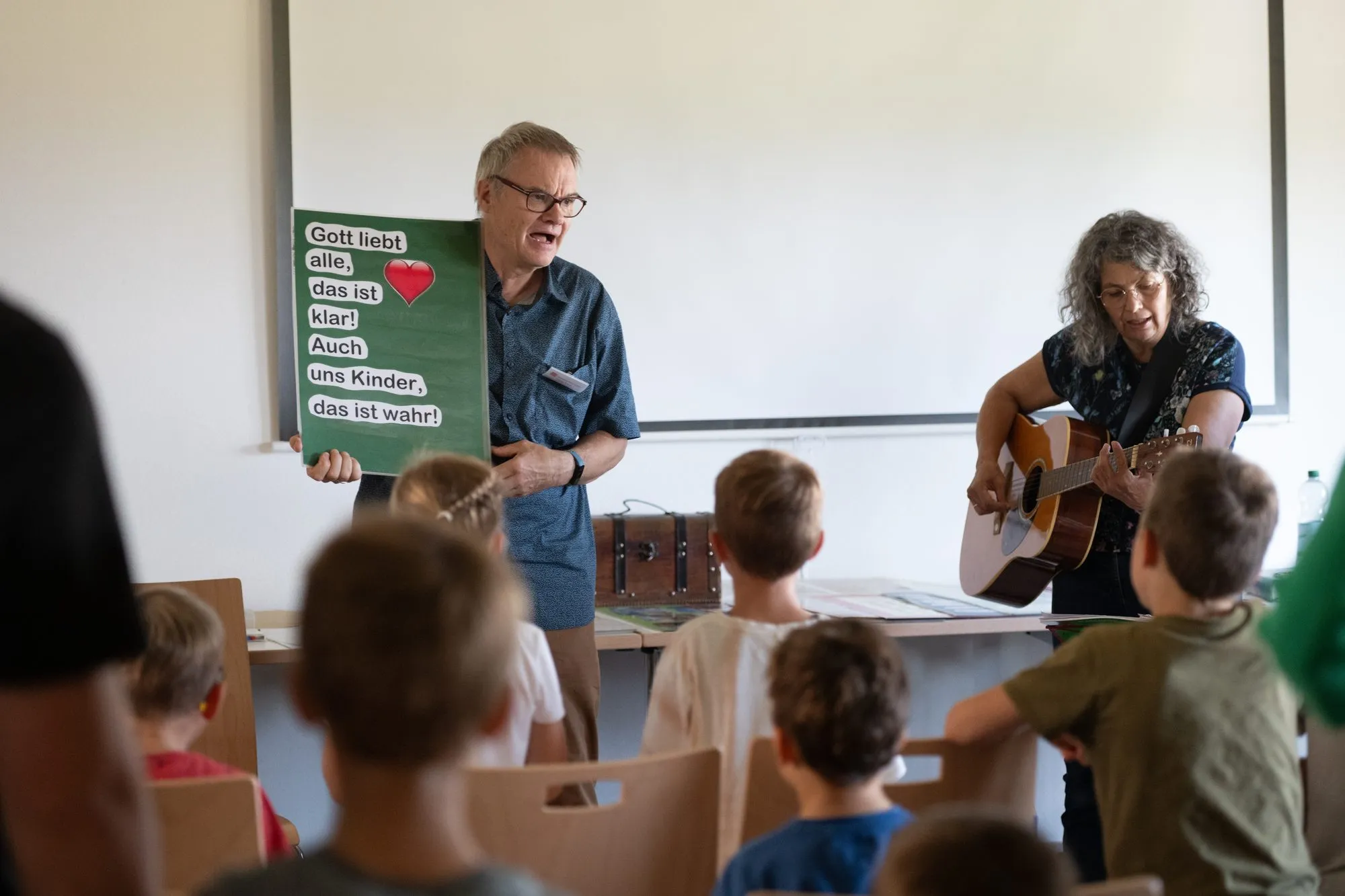 Im Kinderprogramm: ein Mitarbeiter hält ein Plakat „Gott liebt alle“, eine Mitarbeiterin spielt Gitarre, davor sitzen Kinder.