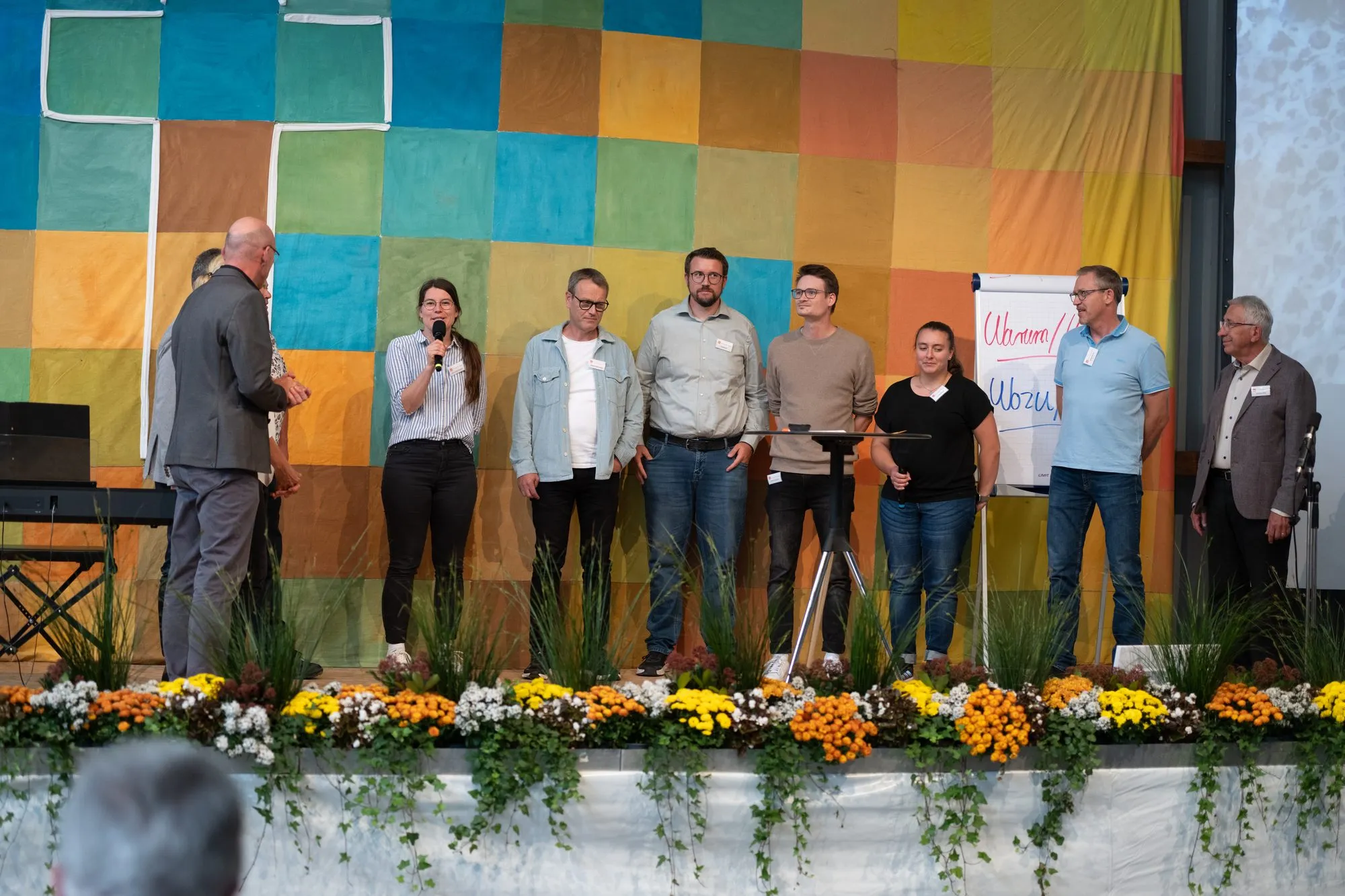 Das Team von Christen helfen Christen steht gemeinsam auf der Bühne vor der bunten Wand, davor Blumenschmuck.
