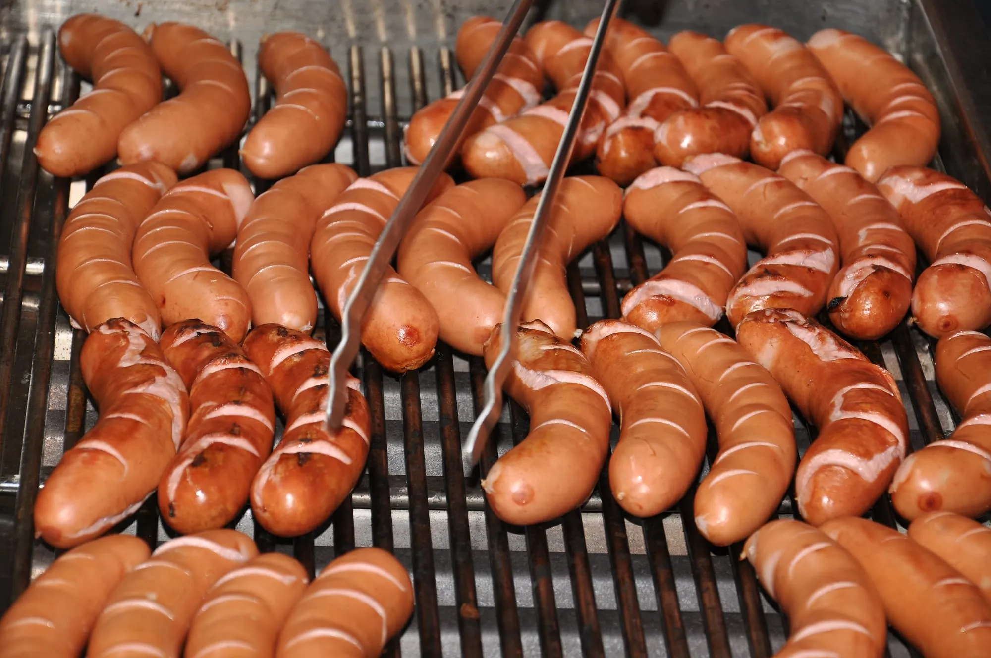 Auf dem Grill liegen dicht an dicht Würste, dazwischen eine Grillzange.