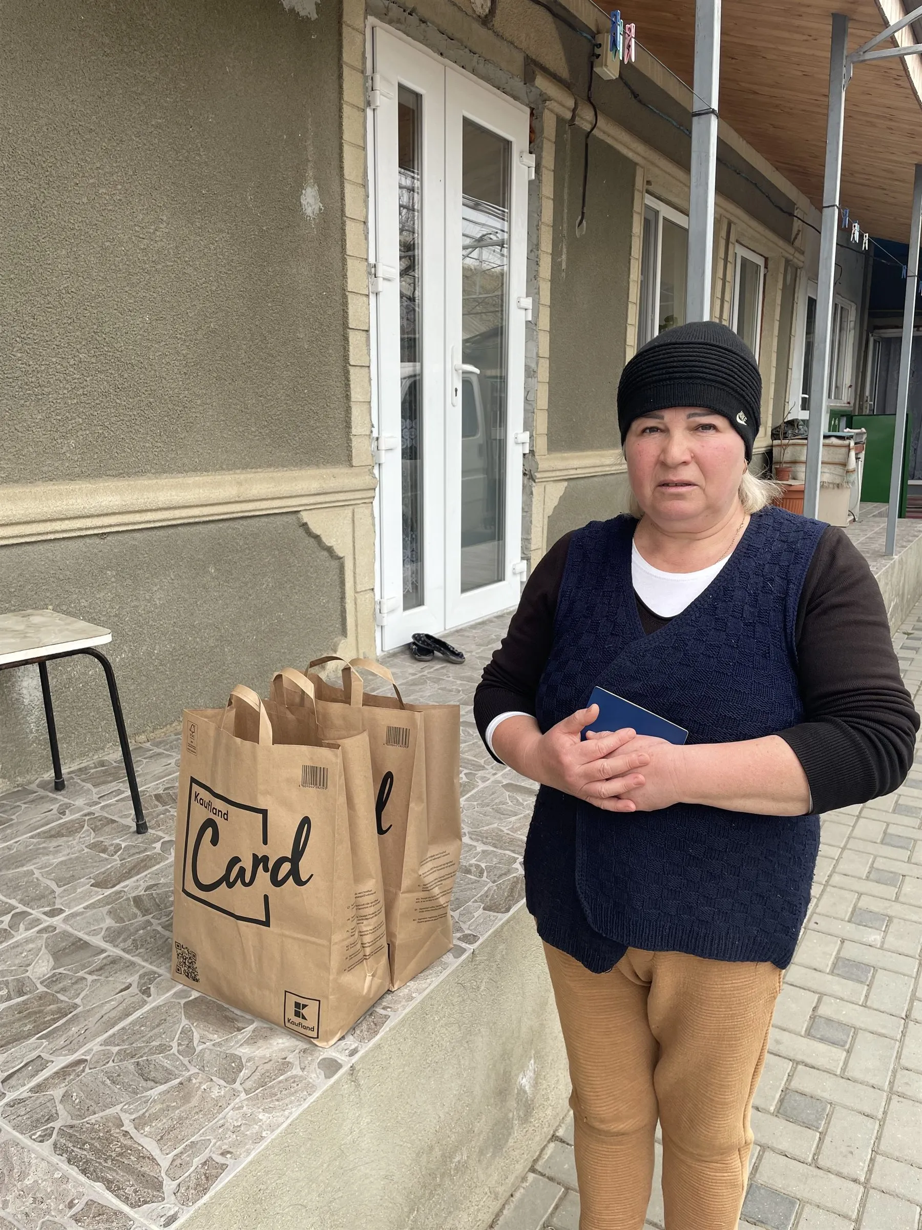 Eine Frau steht vor einem Haus, neben ihr zwei gefüllte Papiertüten.