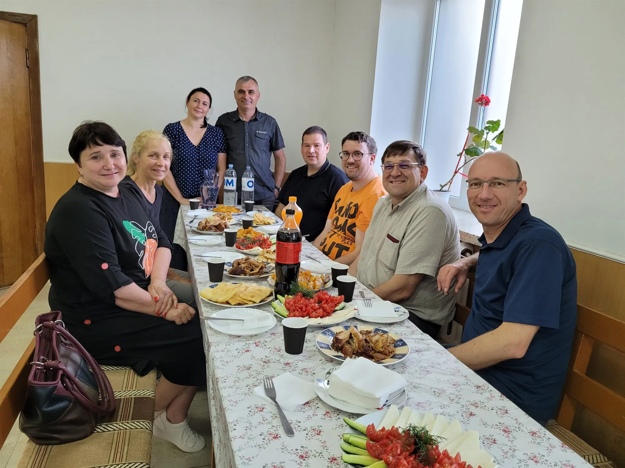 Eine Gruppe sitzt bei einem Besuch an einem lang gedeckten Tisch mit einheimischen Speisen und schaut in die Kamera.