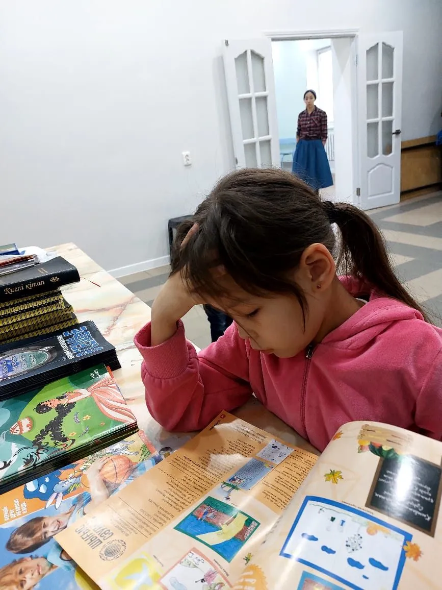Ein Mädchen liest an einem Tisch in einer aufgeschlagenen Kinderzeitschrift, daneben liegt ein Stapel Hefte.